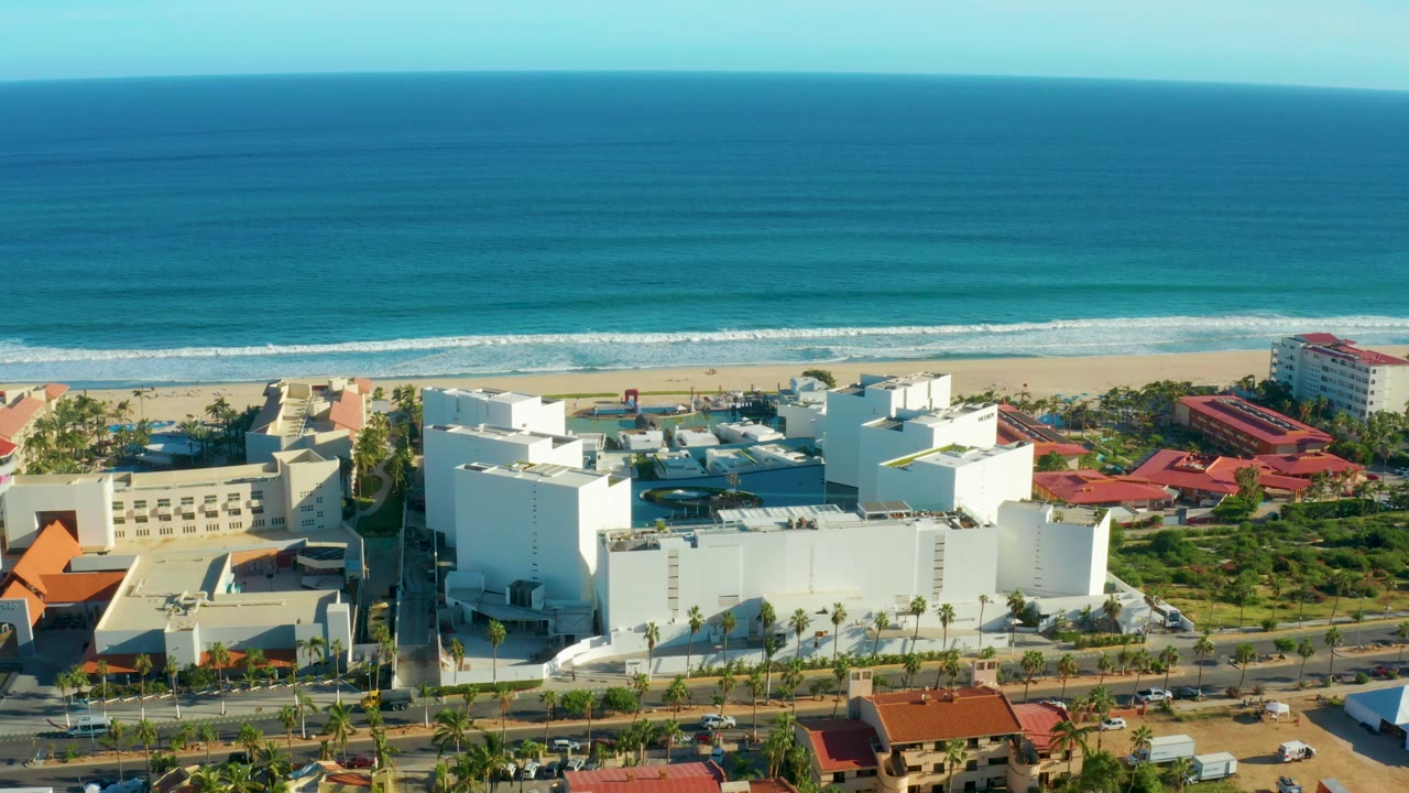 Aerial frame over San Jose Del Cabo showing white coastal buildings, red roofs, a roadside edge, sandy beach, blue ocean, and no visible people.
