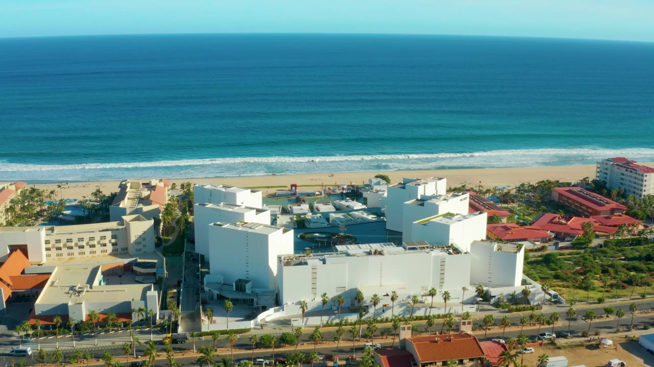 Aerial frame over San Jose Del Cabo showing more of the central white building complex, palm-lined road, narrow beach, layered blue ocean, and no visible people.