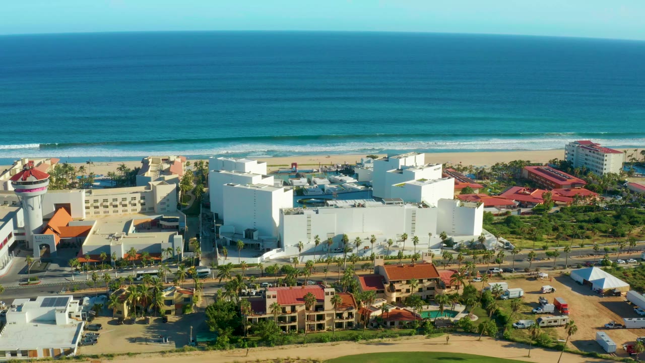 San Jose del Cabo aerial view with a white building complex, roadside cars, palms, red-roofed structures, beach surf, and a broad blue ocean beyond with no visible people.