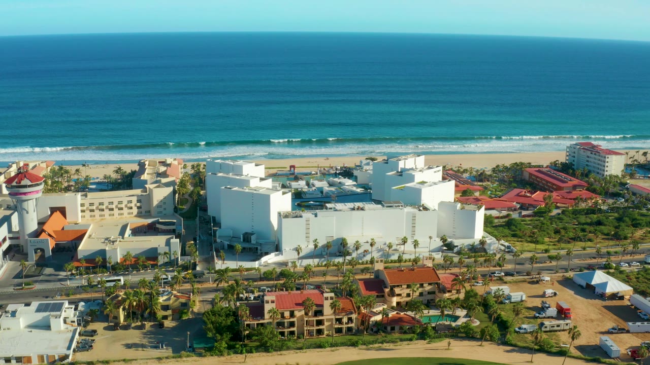 San Jose del Cabo aerial frame with a round-topped resort tower near the left edge, large white buildings, red roofs, road traffic, palms, beach, and blue ocean with no visible people.
