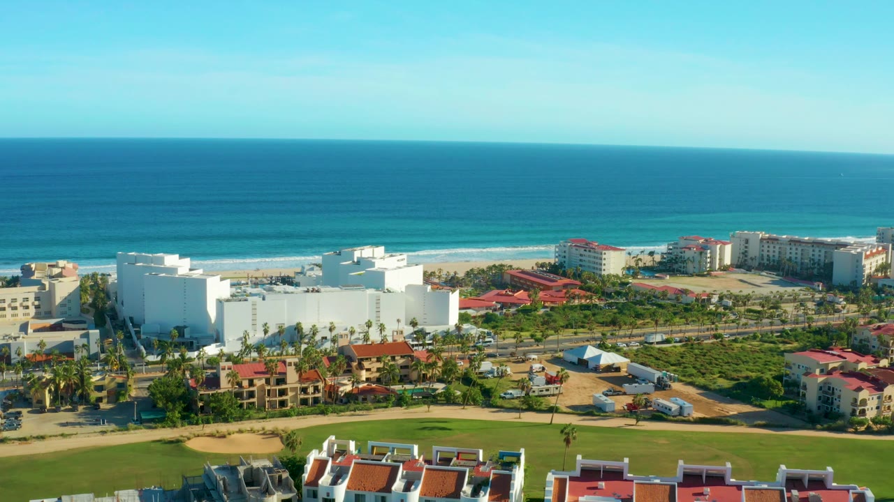 San Jose del Cabo aerial frame with the ocean horizon high in view, beach and surf behind a large white building, red roofs, roads with small vehicles, palms, and no visible people.