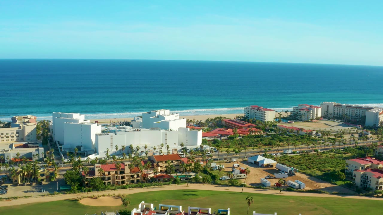 A high drone view in San Jose del Cabo shows the white coastal building in the lower left, red-roofed structures across the midground, beach and ocean beyond, and no visible people.