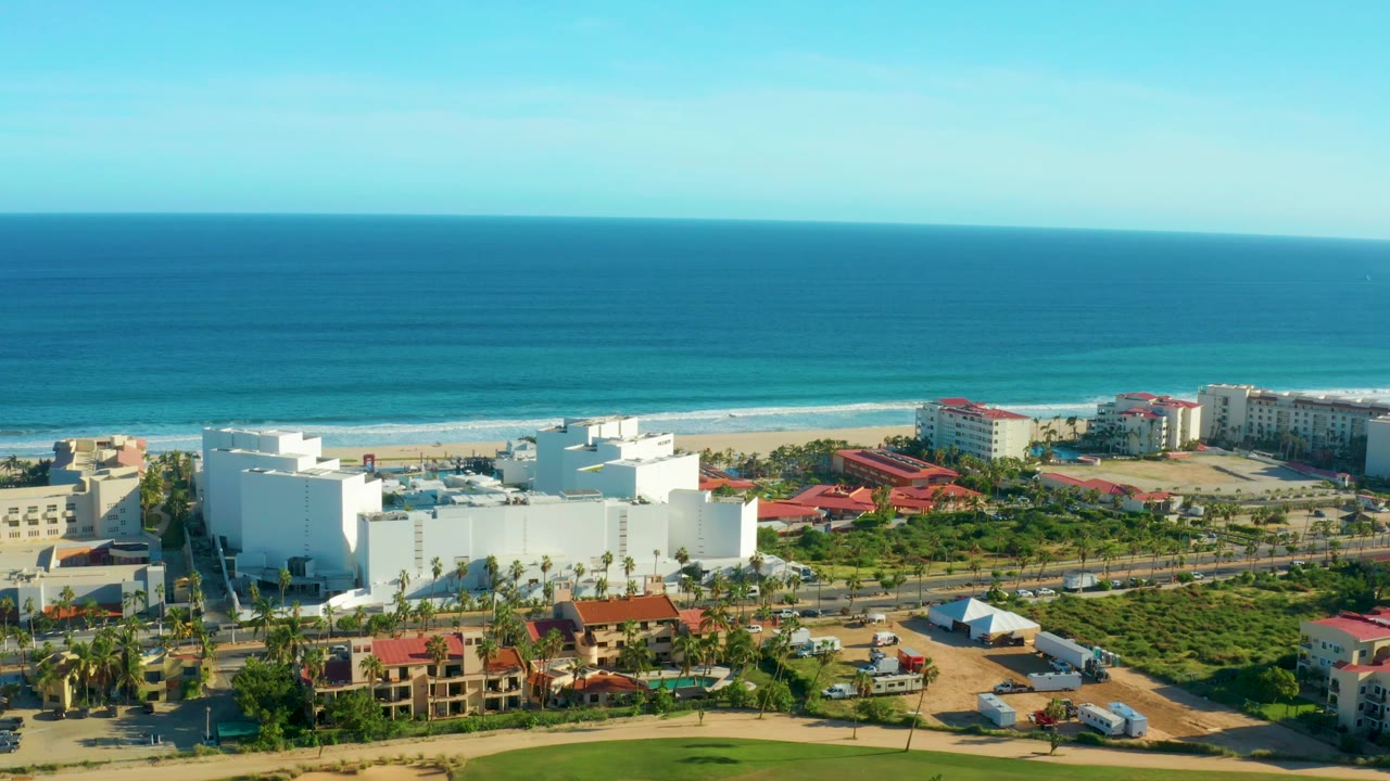 Mid-clip aerial view over San Jose del Cabo with bright blue ocean, thin surf bands, sandy beach, white building mass, nearby red roofs, green areas, roads, vehicles, and no visible people.