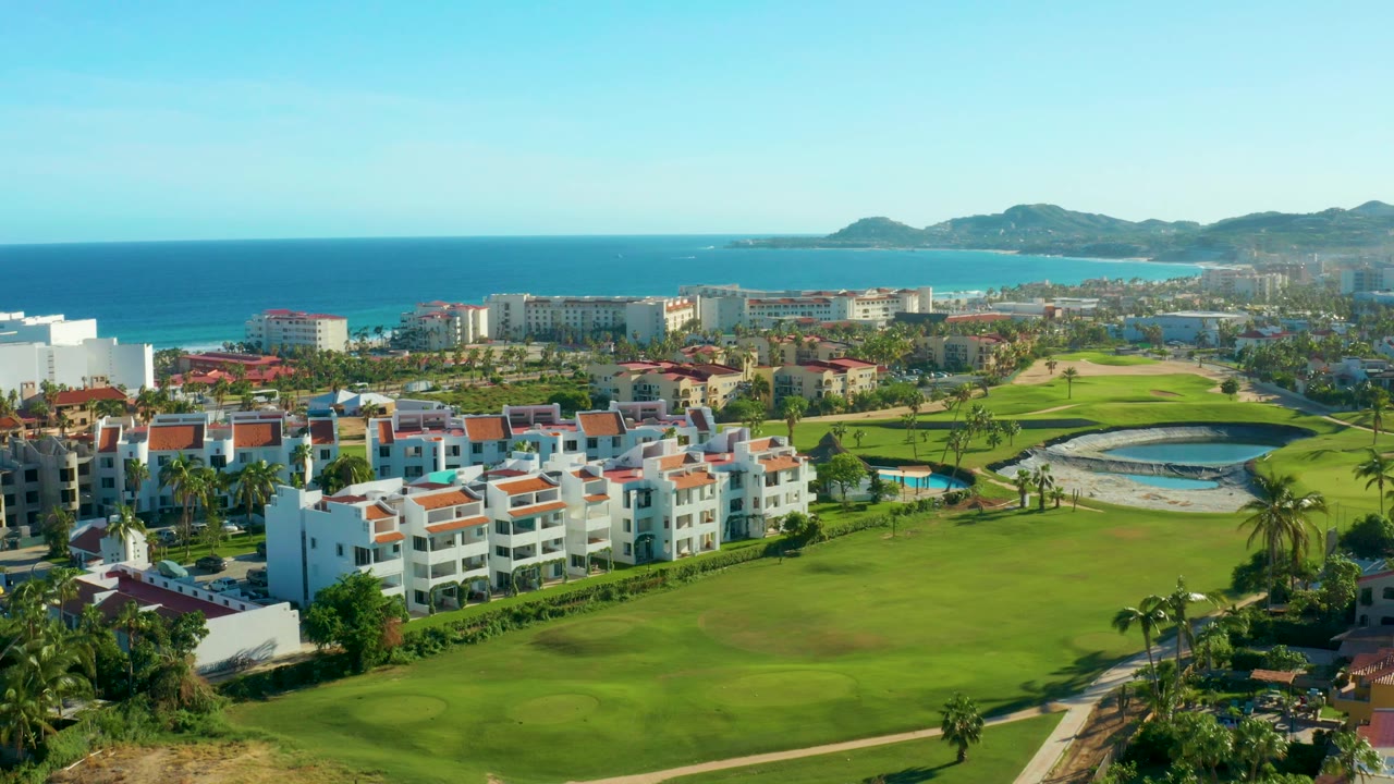 Aerial frame from San Jose del Cabo, Baja California Sur, showing white residential buildings with red roofs, green golf fairways, blue ocean, distant hills, and no visible people.