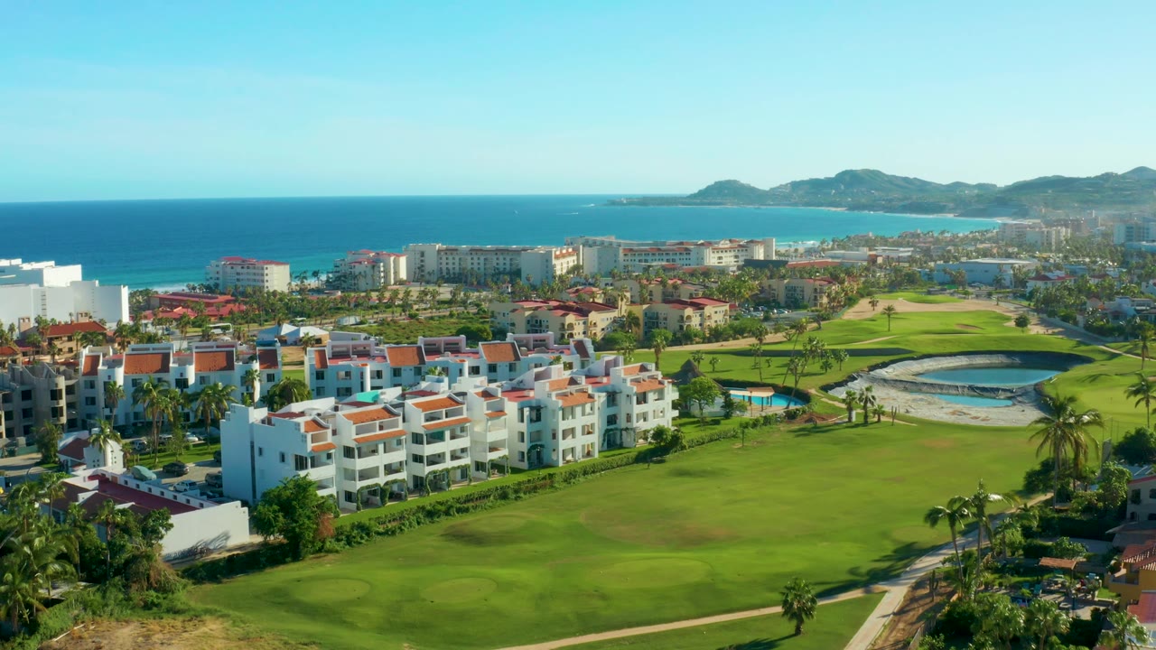 High aerial view over red-roofed buildings, fairway grass, beach, and blue sea.