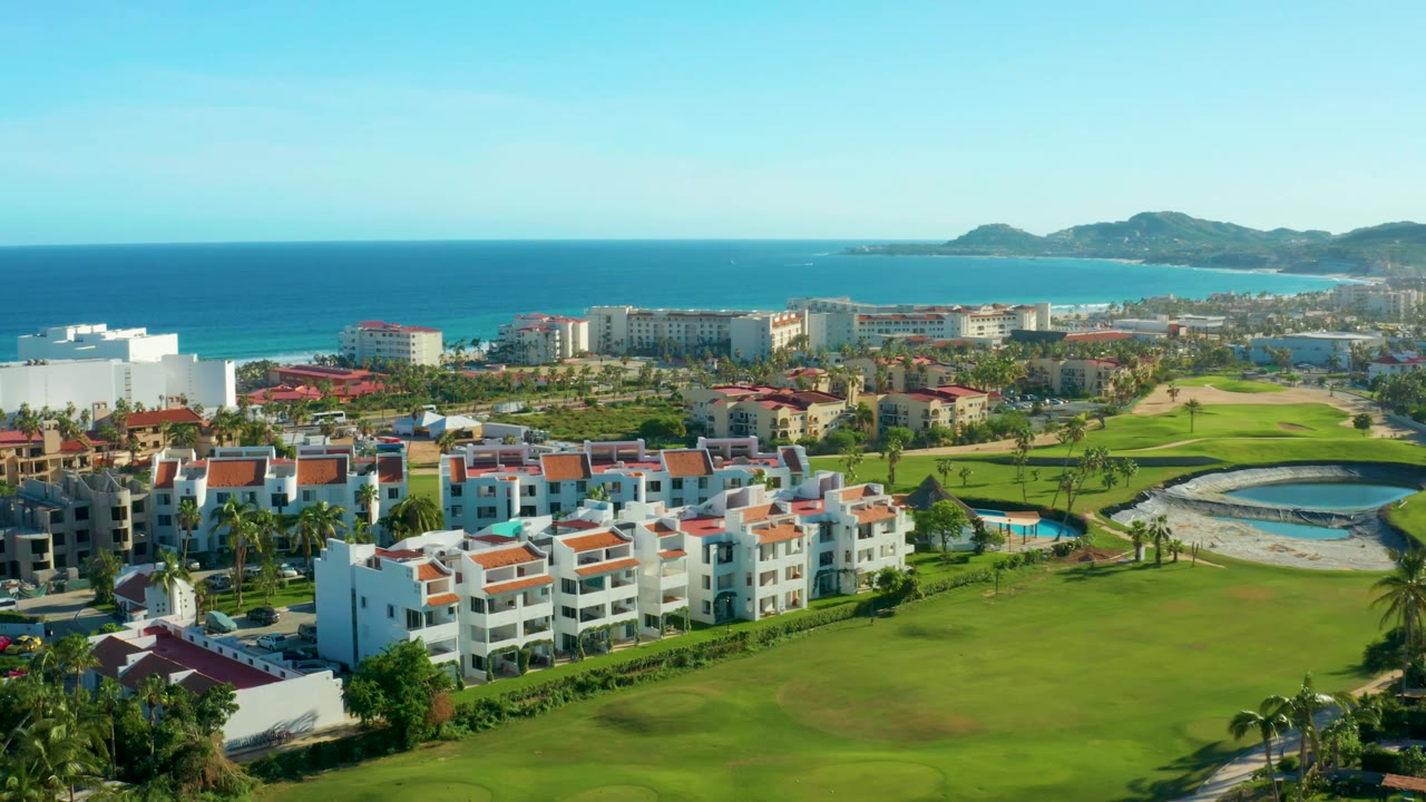 San Jose del Cabo aerial frame showing white apartment buildings, a green golf course with a curved water feature, beachfront structures, blue water, distant hills, and no visible people.