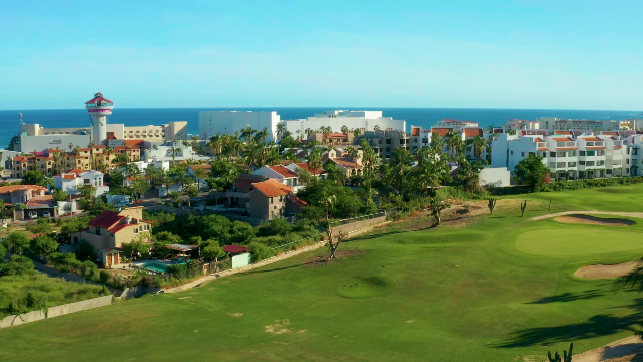 San Jose del Cabo aerial frame with golf fairway and sand bunker in front of residential buildings, a lighthouse-like tower at left, blue ocean behind, and no visible people.