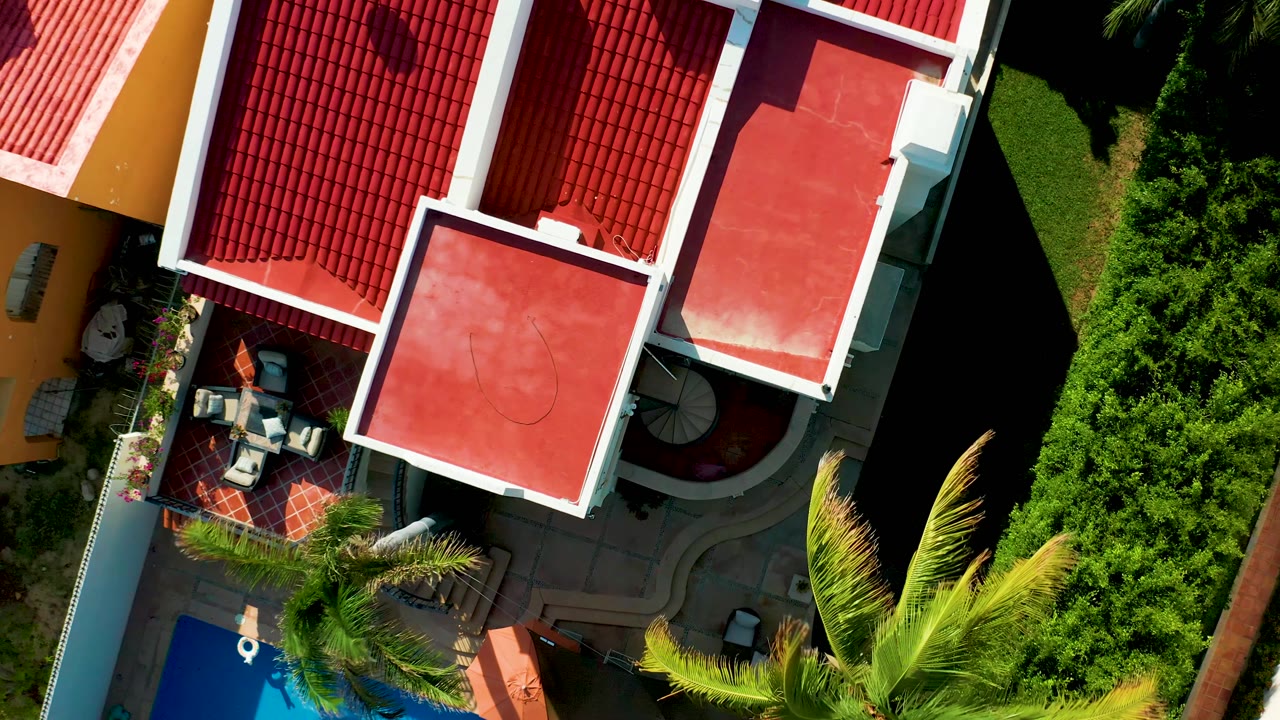 Overhead San Jose Del Cabo frame showing red roof planes, patio chairs under shade, a visible spiral stair shape, pool blue near the lower edge, palm fronds, hedges, and no visible people.