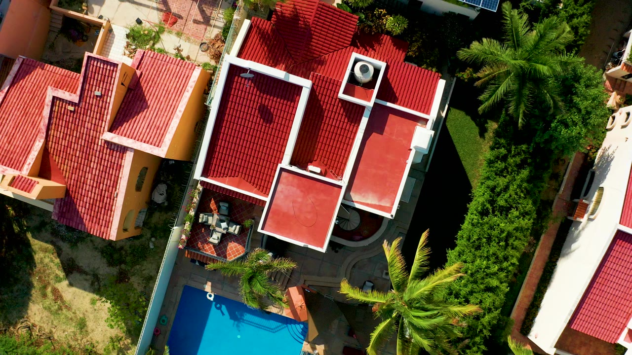Overhead frame in San Jose del Cabo showing red tile roofs, a blue pool corner, palm trees, green hedges, and enclosed patios with no visible people.