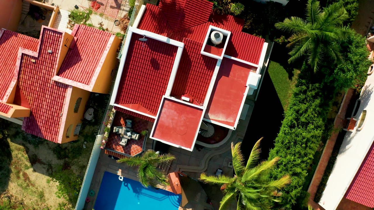 Overhead San Jose del Cabo frame centered on red roofs and white roof trim, with palms and a blue pool corner along the lower edge and no visible people.