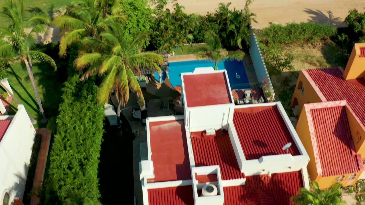 Overhead aerial frame in San Jose Del Cabo showing red-tiled residential roofs, white walls, palm trees, dense greenery, a blue pool, and sandy beach edge, with no visible people.