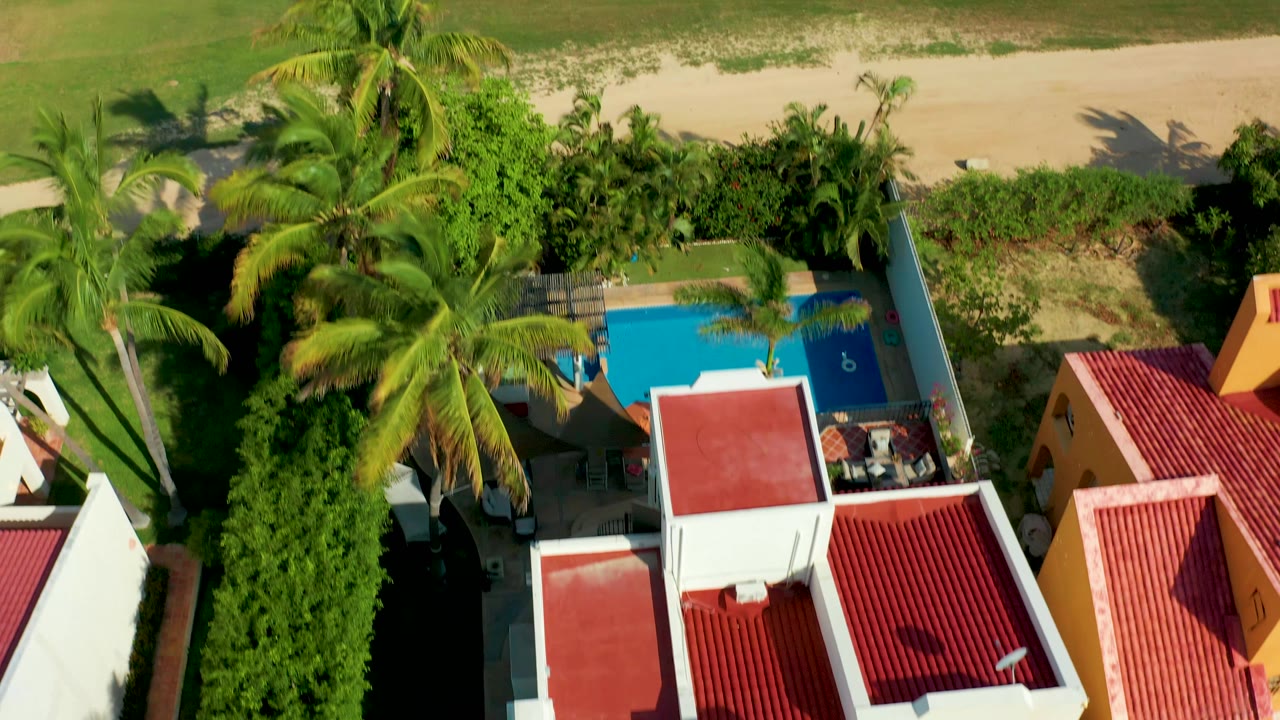 Overhead San Jose Del Cabo frame showing palm crowns crossing above a blue pool, red tile roofs, a yellow building, hedges, and the sandy beach edge beyond, with no visible people.