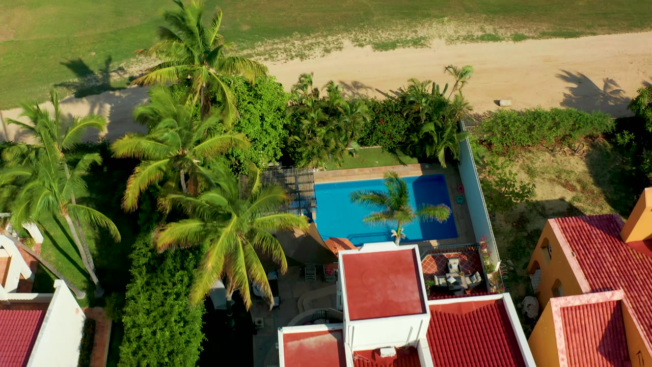 Top-down frame with palms, dense hedges, pool corner, red roofs, and beach sand.