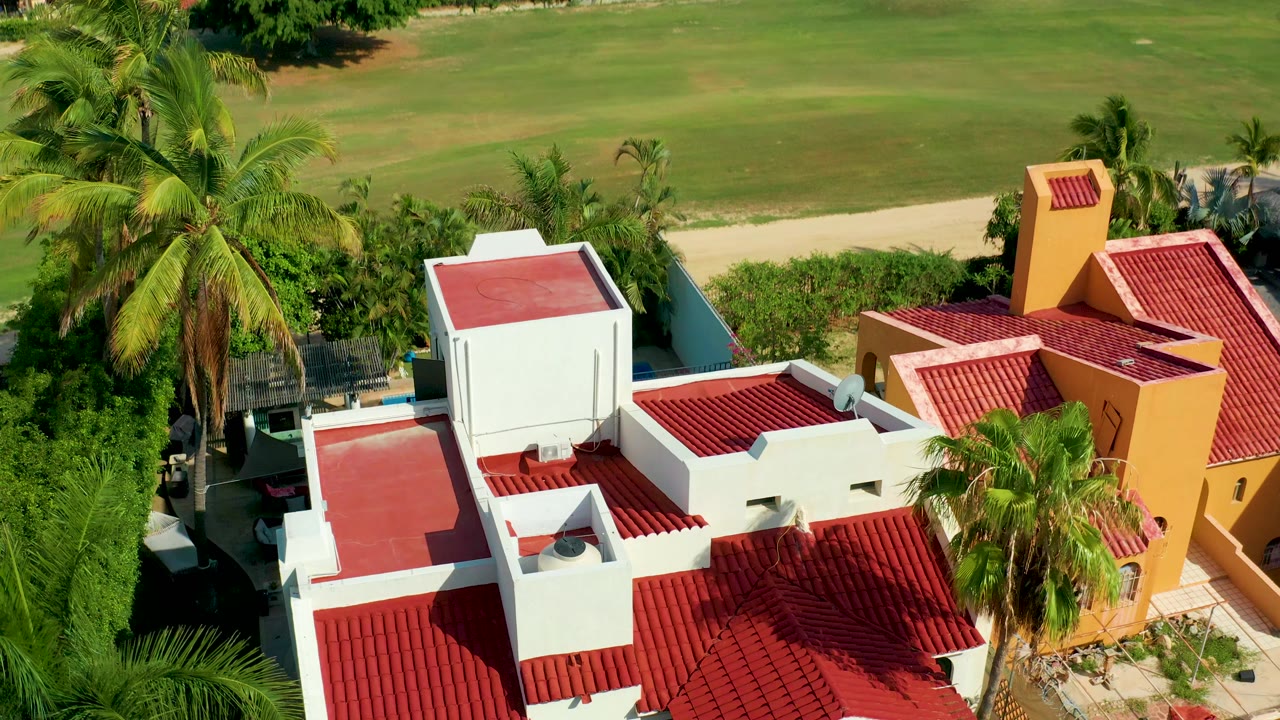 Overhead drone frame in San Jose del Cabo showing white and yellow homes with red roofs, tall palms, a hedge line, and a green field beyond the property edge, with no visible people.