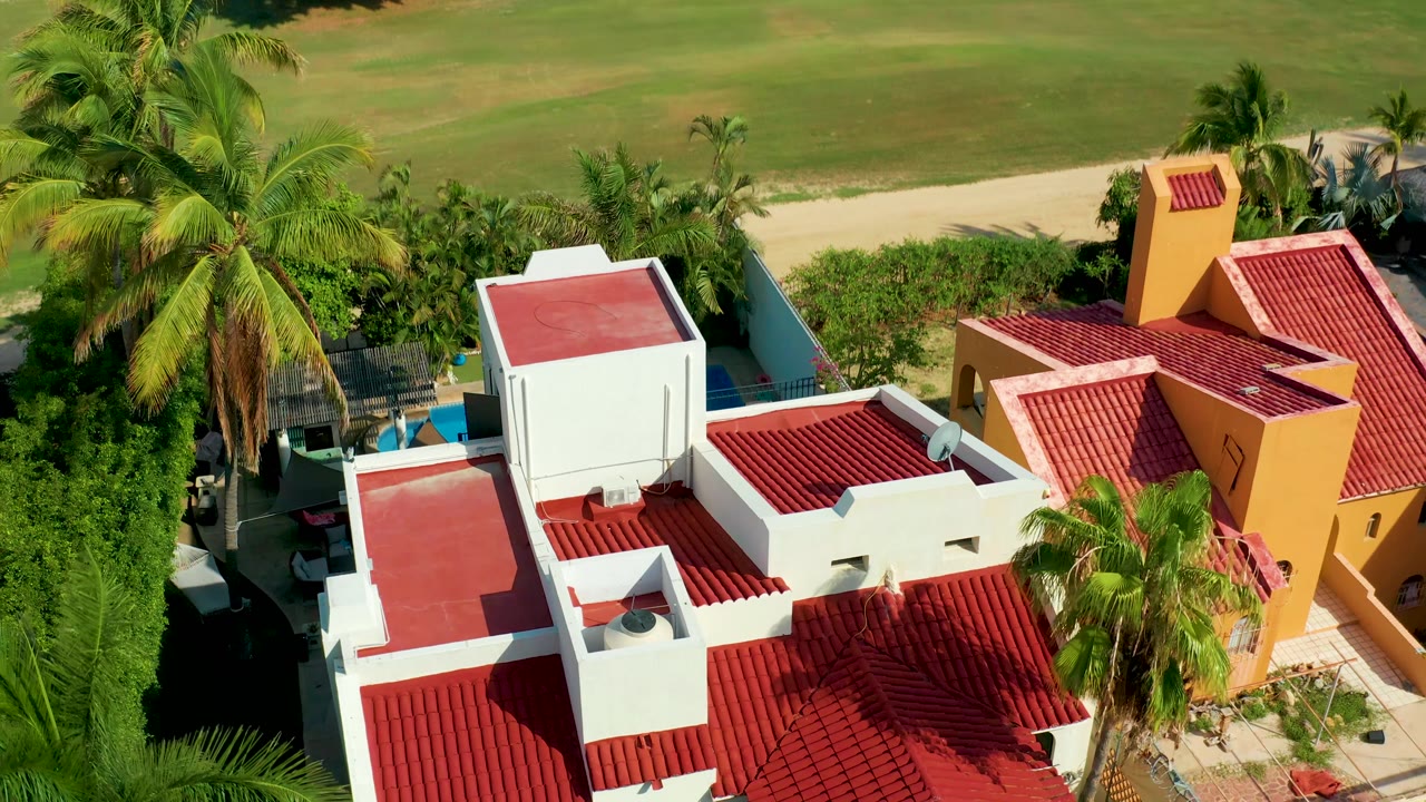 Overhead frame in San Jose del Cabo with red-roofed residential buildings, palms, hedges, a field beyond a dirt path, and a small section of blue swimming pool beginning to appear.