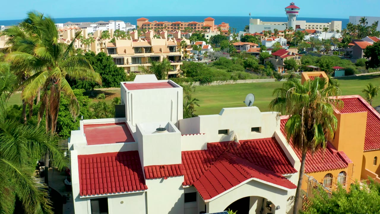 Opening aerial frame over San Jose del Cabo homes with red tiled roofs in front, palm trees around them, a green lawn beyond, distant buildings, ocean, and no visible people.