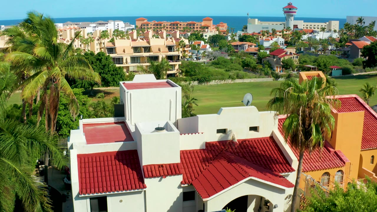 Early aerial frame in San Jose del Cabo showing a white rooftop volume, red tile roof sections, palms at both sides, a broad lawn, distant buildings, ocean, and no visible people.