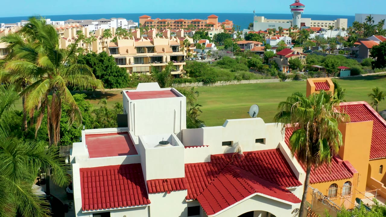 San Jose del Cabo aerial view with the red tiled roof lower in frame, palms bordering the homes, green lawn and residential blocks behind, a narrow ocean band, and no visible people.