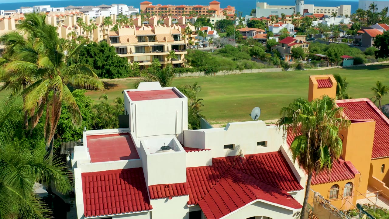 Lower aerial view over red roofs, palms, and a green lawn.