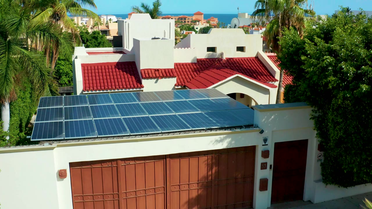 Opening aerial frame in San Jose Del Cabo shows rooftop solar panels in front of a white house with red tile roofing, palms on both sides, ocean on the horizon, and no visible people.