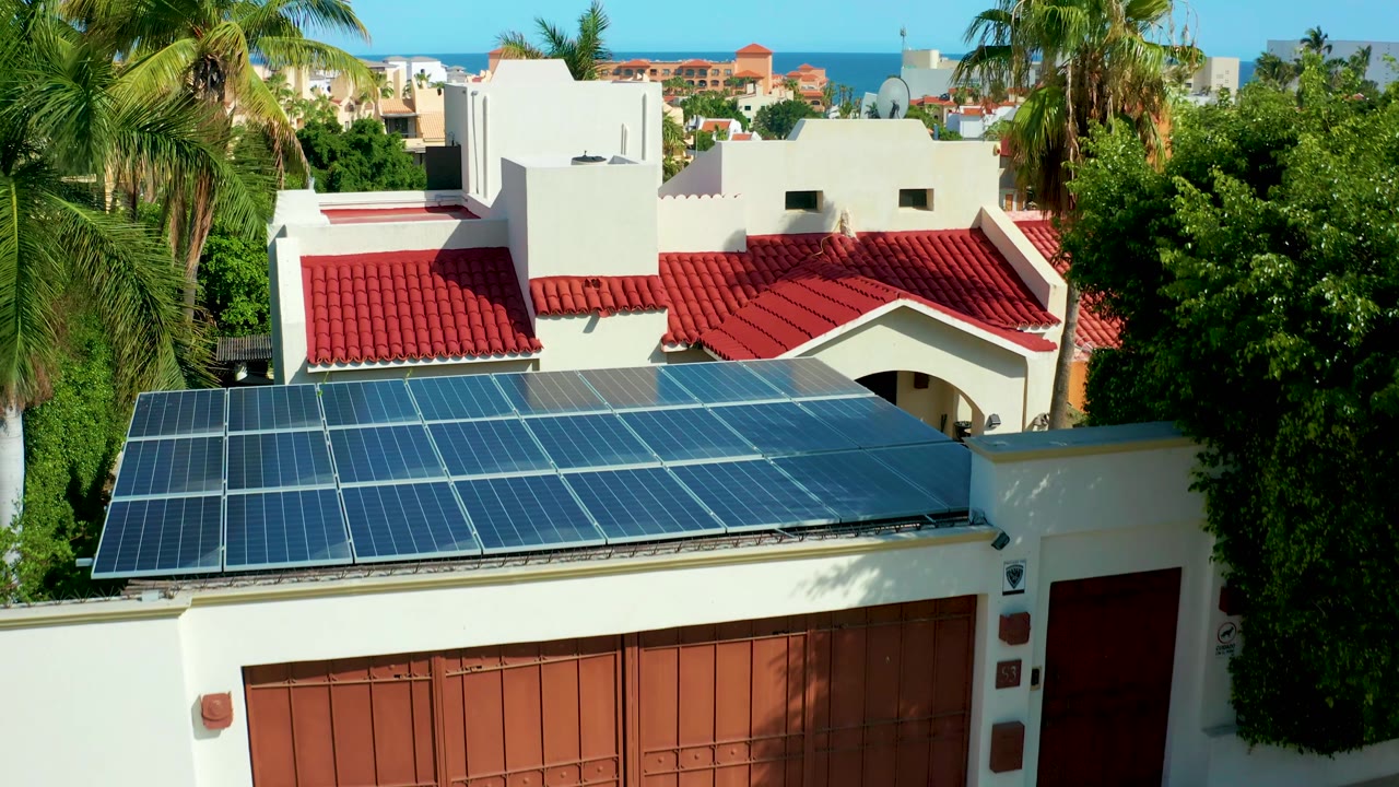 At 0:02, San Jose Del Cabo appears from above with the solar roof angled across the bottom of frame, a white and red-roofed house behind it, palms, distant homes, ocean, and no visible people.