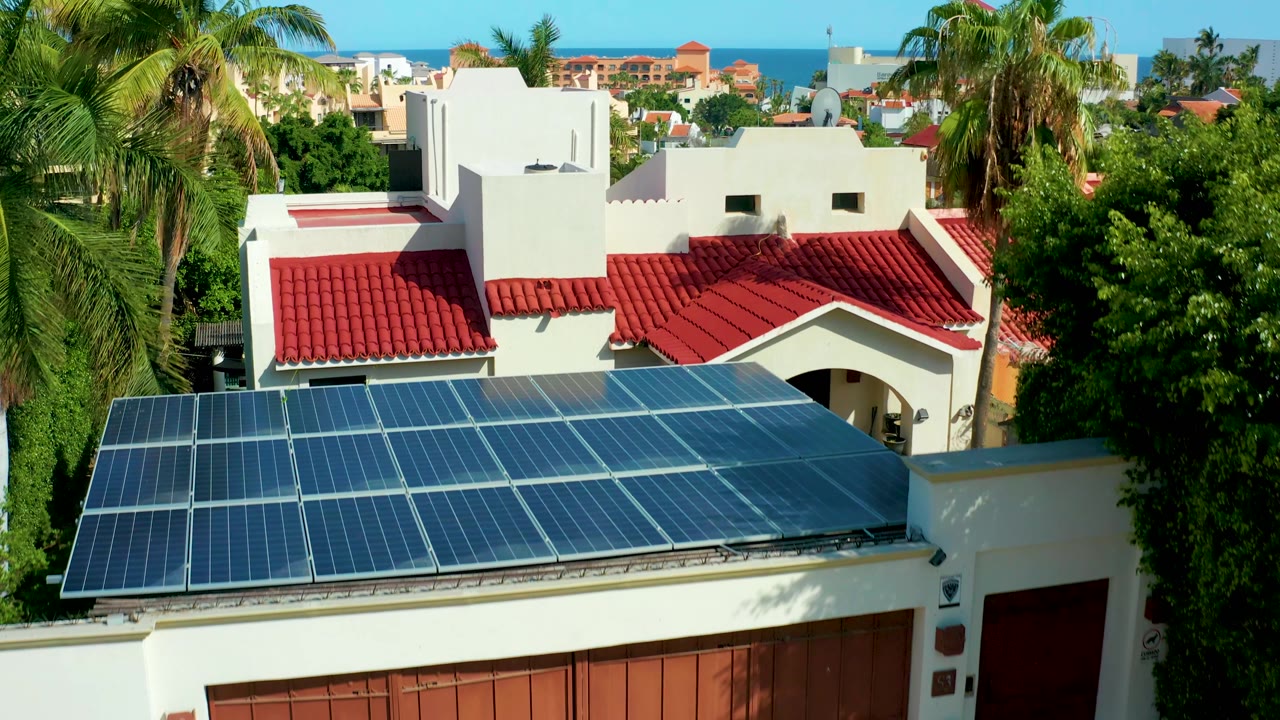 Closer aerial angle on solar panels, red roof, white walls, palms, and ocean horizon.