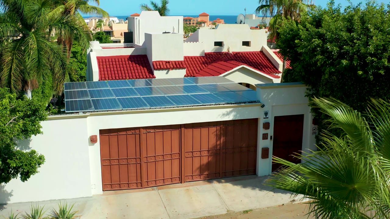 The San Jose Del Cabo residential exterior remains centered, with blue solar panels spanning the wall above the red garage door, palms at the edges, and no visible people.