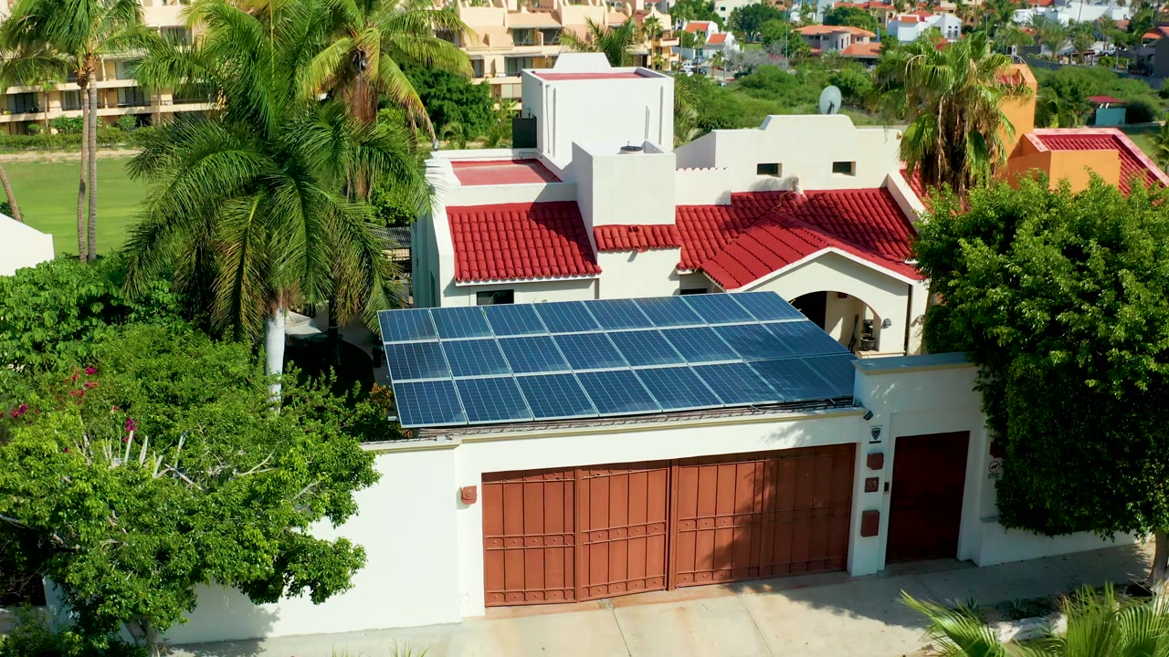 Aerial-style frame of the San Jose Del Cabo house with blue solar panels above brown garage doors, red roof tiles behind them, palms on both sides, background residences, and no visible people.