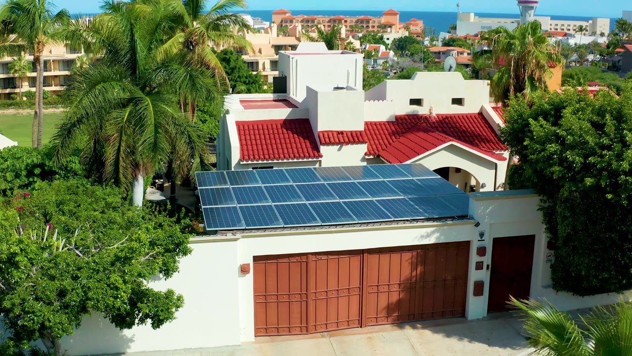 House with rooftop solar panels, palms, greenery, distant ocean, and blue sky.