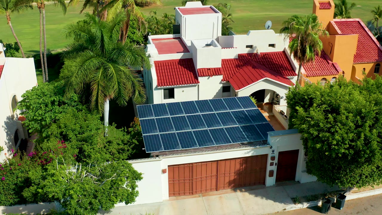 Aerial frame from San Jose del Cabo, Mexico showing a white house with blue solar panels on the garage roof, red tile roofs, surrounding palm trees, and no visible people.