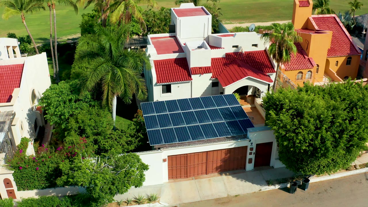 Aerial residential house with solar panels, red roofs, palms, and greenery.