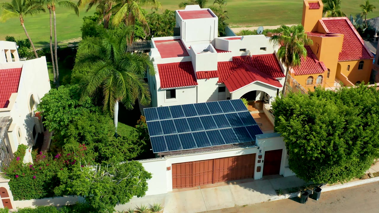 San Jose del Cabo aerial frame showing a white home, orange neighboring structure, red tile roofs, a broad solar-panel array over the garage, palms, and no clearly visible person.
