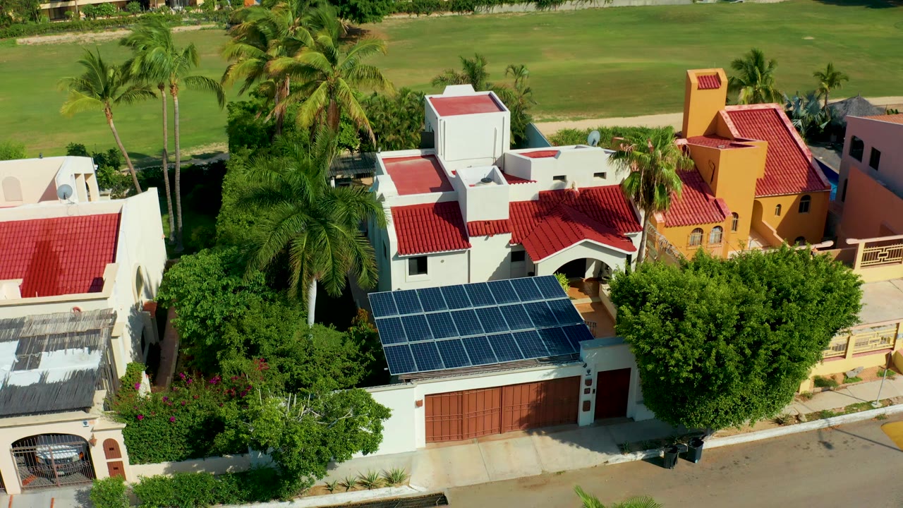 Second frame over San Jose del Cabo homes where the solar panel array sits near the lower edge, palms and lawns fill the upper background, and no visible people appear.