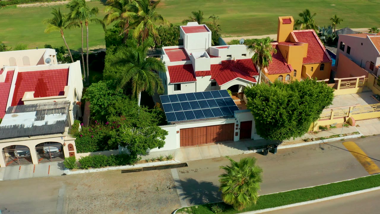 Oblique aerial frame over San Jose del Cabo houses with red roofs, a solar panel roof, palm trees, a road edge, and a green field behind them, with no visible people.