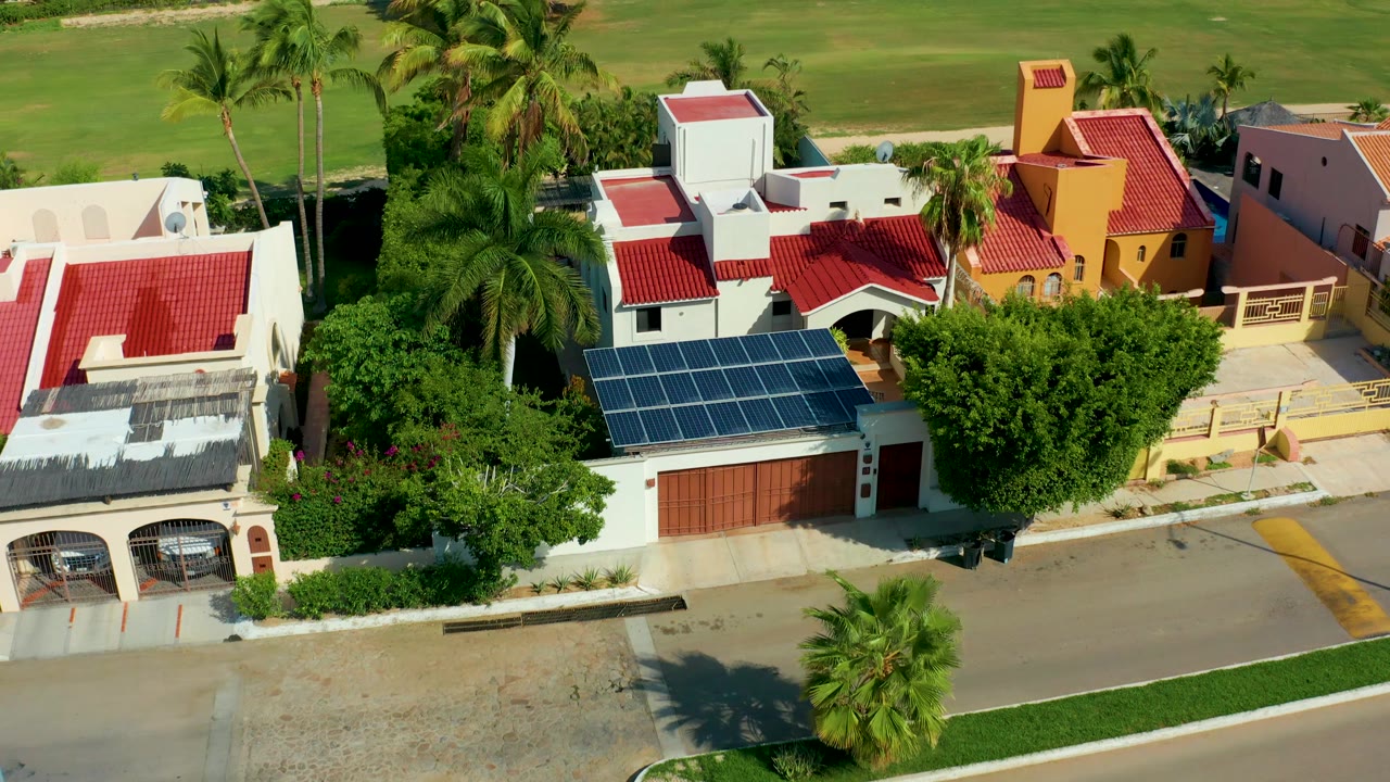 Drone view of colorful residential rooftops, solar panels, palms, and grassland.