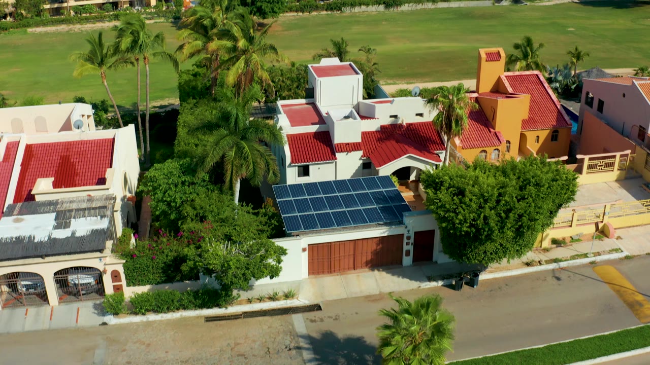 San Jose del Cabo residential aerial with a white house at right, red rooflines across the center, solar panels in the lower left, palms, and a broad green field in the background, with no visible people.