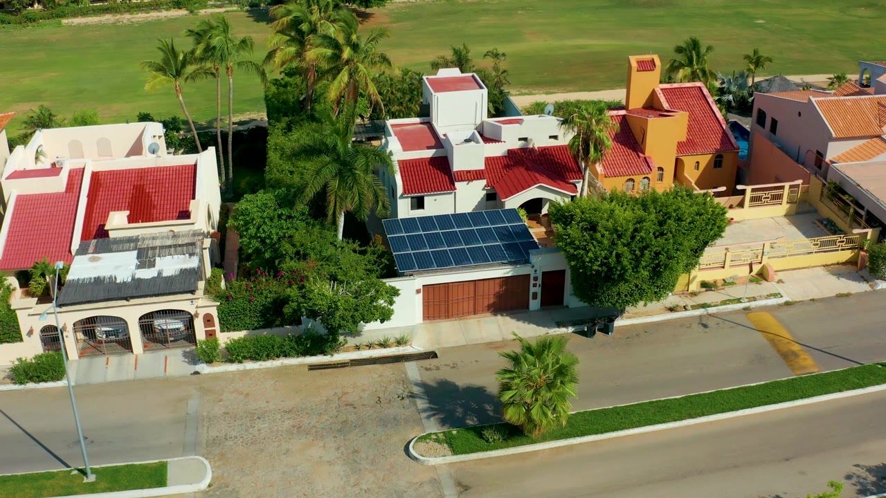 Elevated San Jose del Cabo neighborhood view with the solar-panel roof centered lower in frame, red tile roofs behind it, palm trees at left and right, and no visible people.