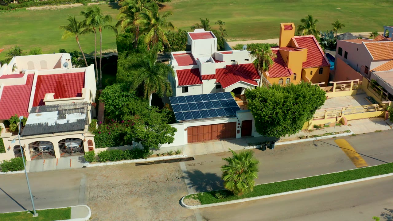 Aerial view in San Jose del Cabo showing the residential street, a white home with solar panels, red-roofed neighboring houses, green open land in back, and no visible people.