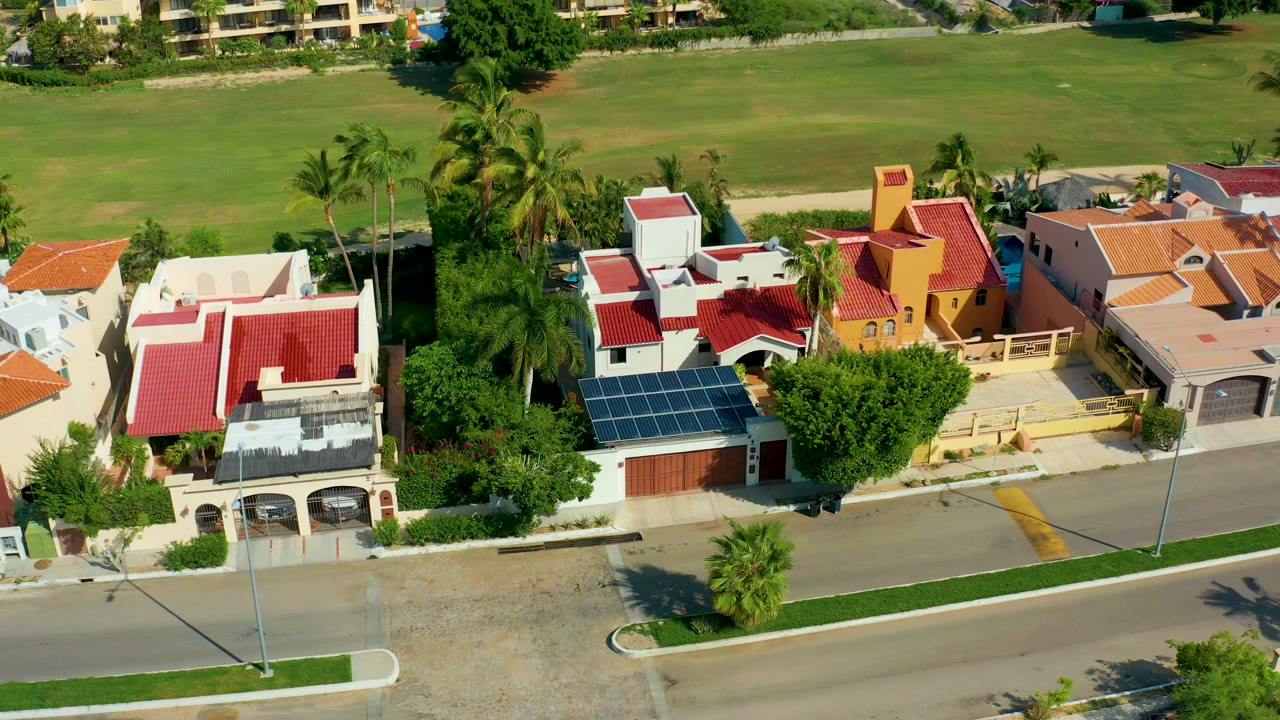 Opening aerial frame over San Jose Del Cabo homes with red roofs, rooftop solar panels, palm trees, a paved street, and a grassy field beyond, with no visible people.