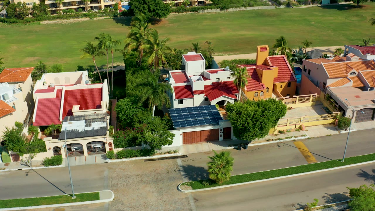 San Jose Del Cabo houses with red, white, yellow, and tan facades sit below the drone, with palms and a broad green field behind them and no visible people.
