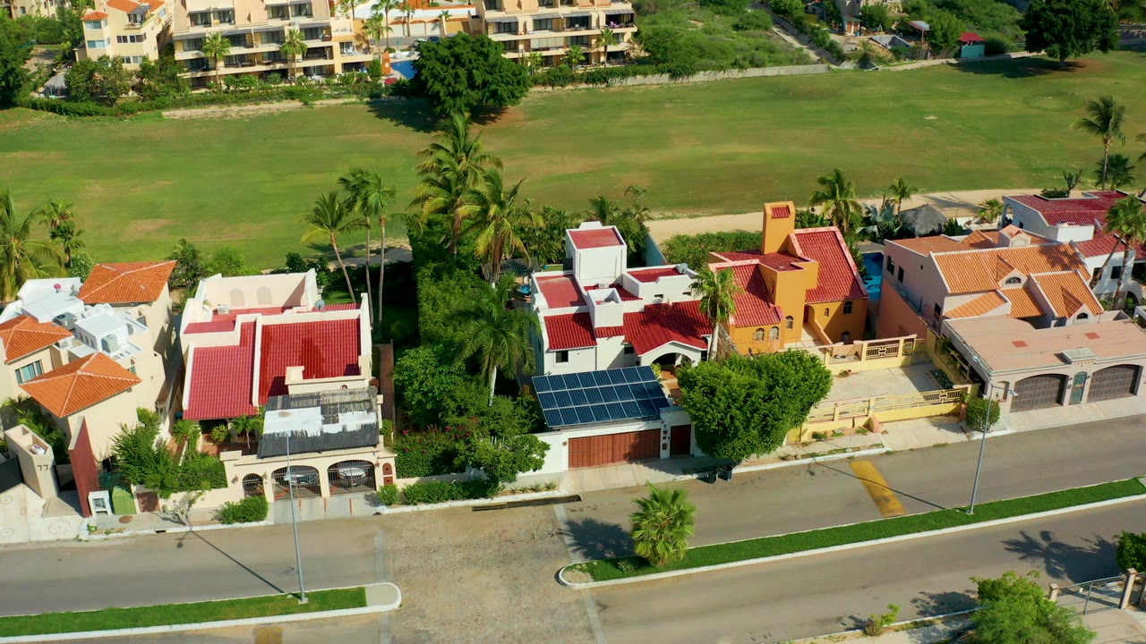 Opening aerial frame over San Jose Del Cabo homes with red and orange roofs, a paved street in front, palms between houses, a green field behind, and no visible people.