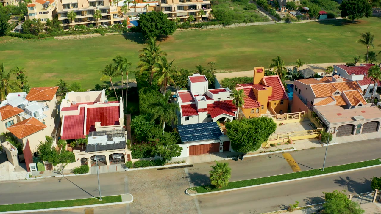 Early aerial view in San Jose Del Cabo with a red-roofed house near the lower left, a yellow house near center, tall palms, a street edge, green field, and no visible people.