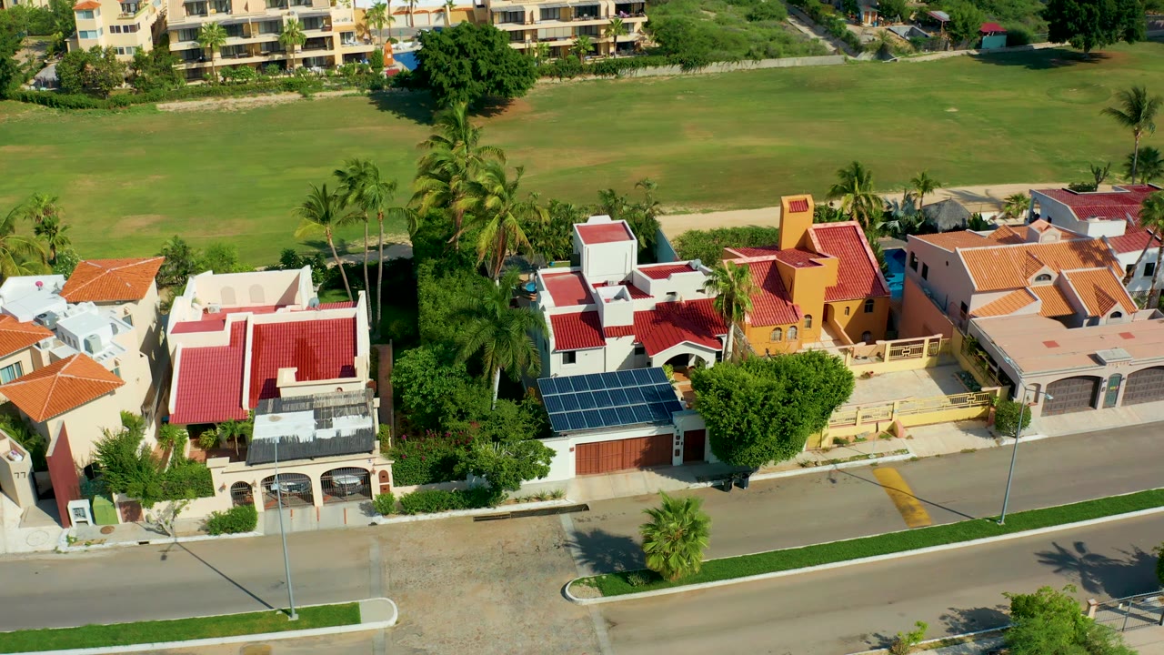 Aerial neighborhood view with palms, rooflines, road, and green field.