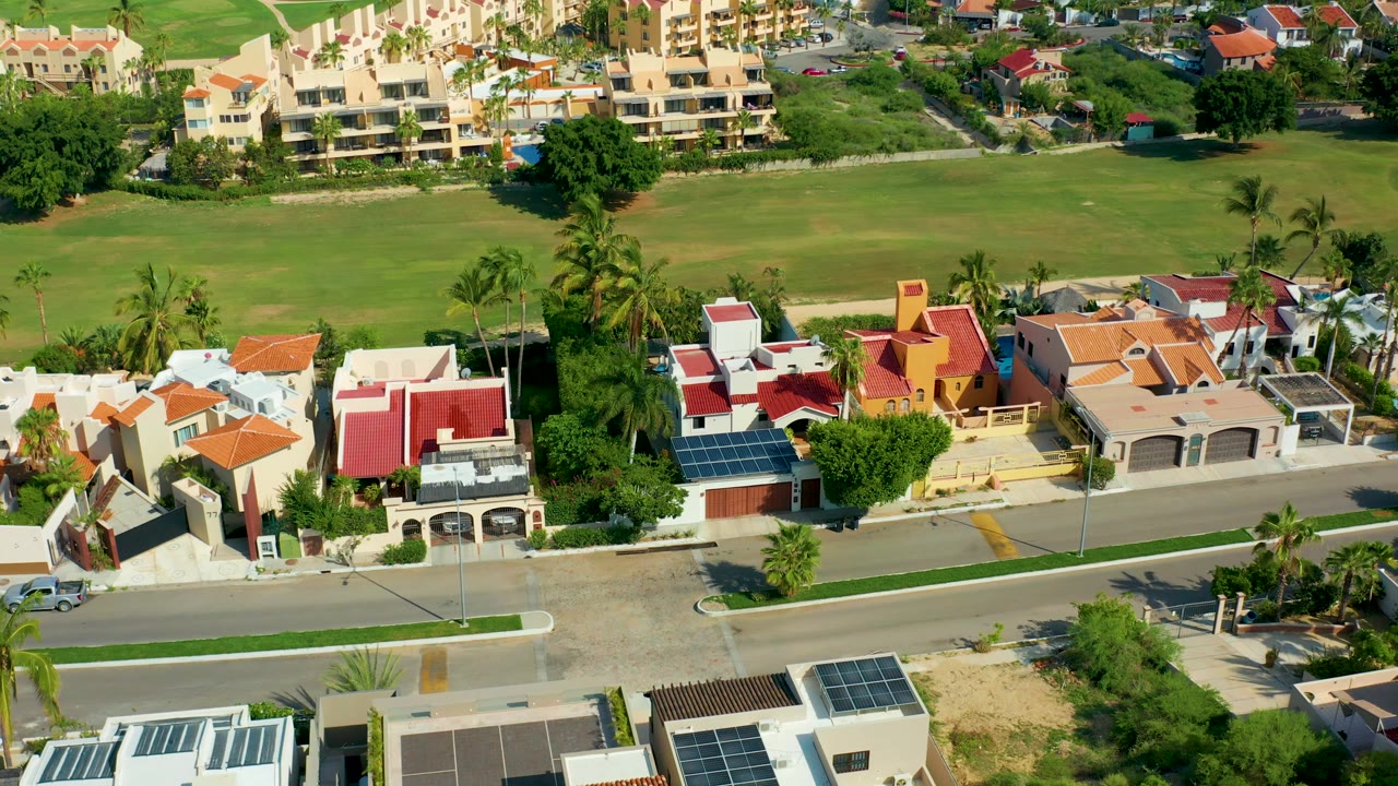 Elevated neighborhood view with solar rooftops, palms, road, and field.