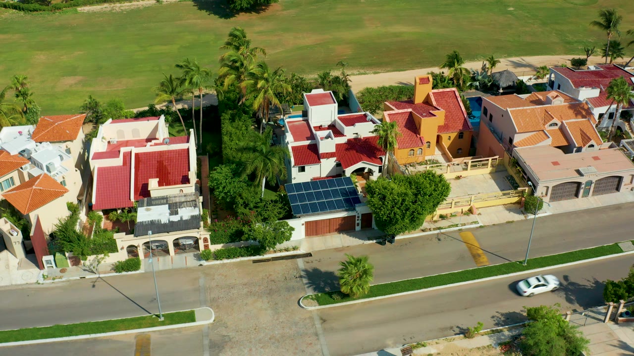 Aerial frame over San Jose del Cabo homes with red and orange roofs, palms, solar panels, a road at the bottom, a green field behind the block, and no visible people.
