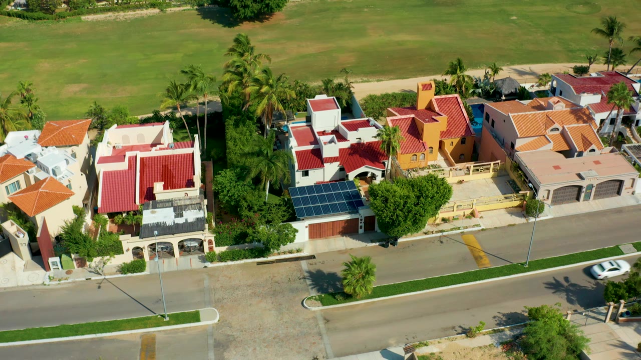 San Jose del Cabo aerial frame with the white car farther along the road, red-roofed houses and solar panels above it, palms between homes, a grassy field in back, and no visible people.