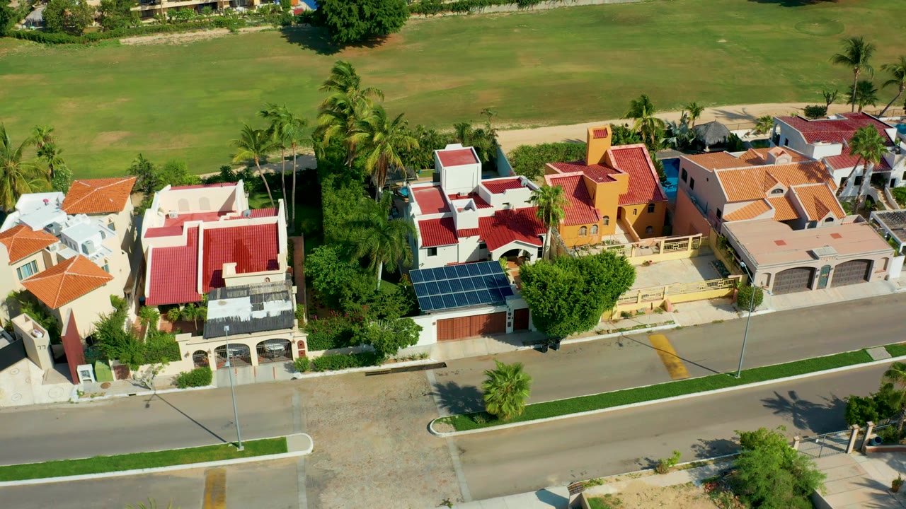 Aerial neighborhood view with homes, palms, road, and green field.