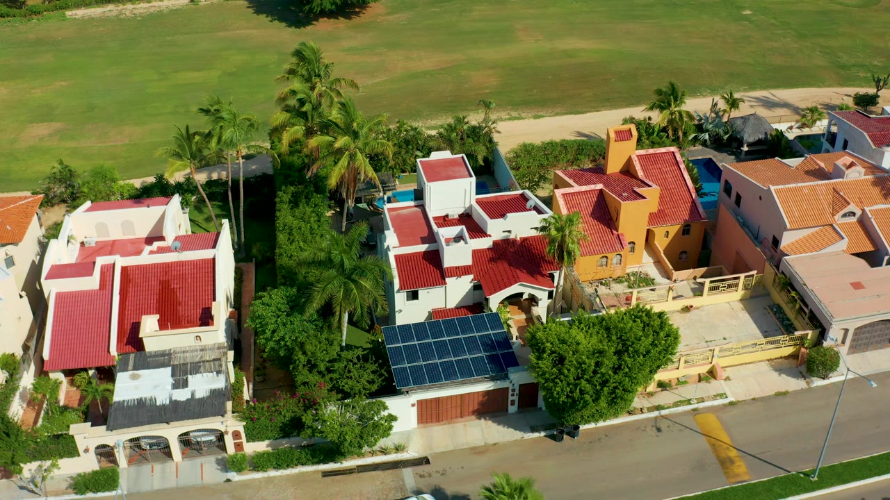 Aerial frame over San Jose Del Cabo homes with red roofs, palm trees, solar panels, a paved street, and no visible people.