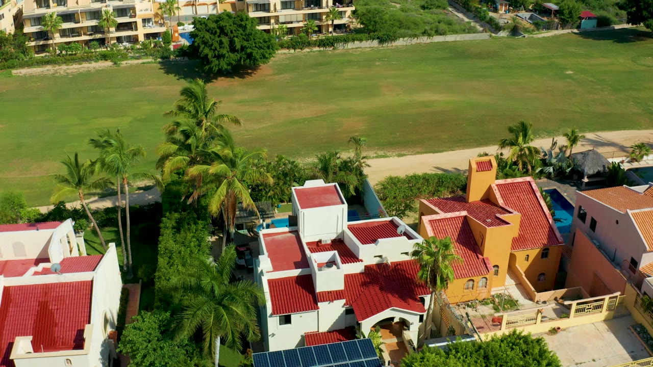 Aerial frame over San Jose Del Cabo homes with red tile roofs, palms, solar panels, a driveway, and a grassy field behind, with no visible people.