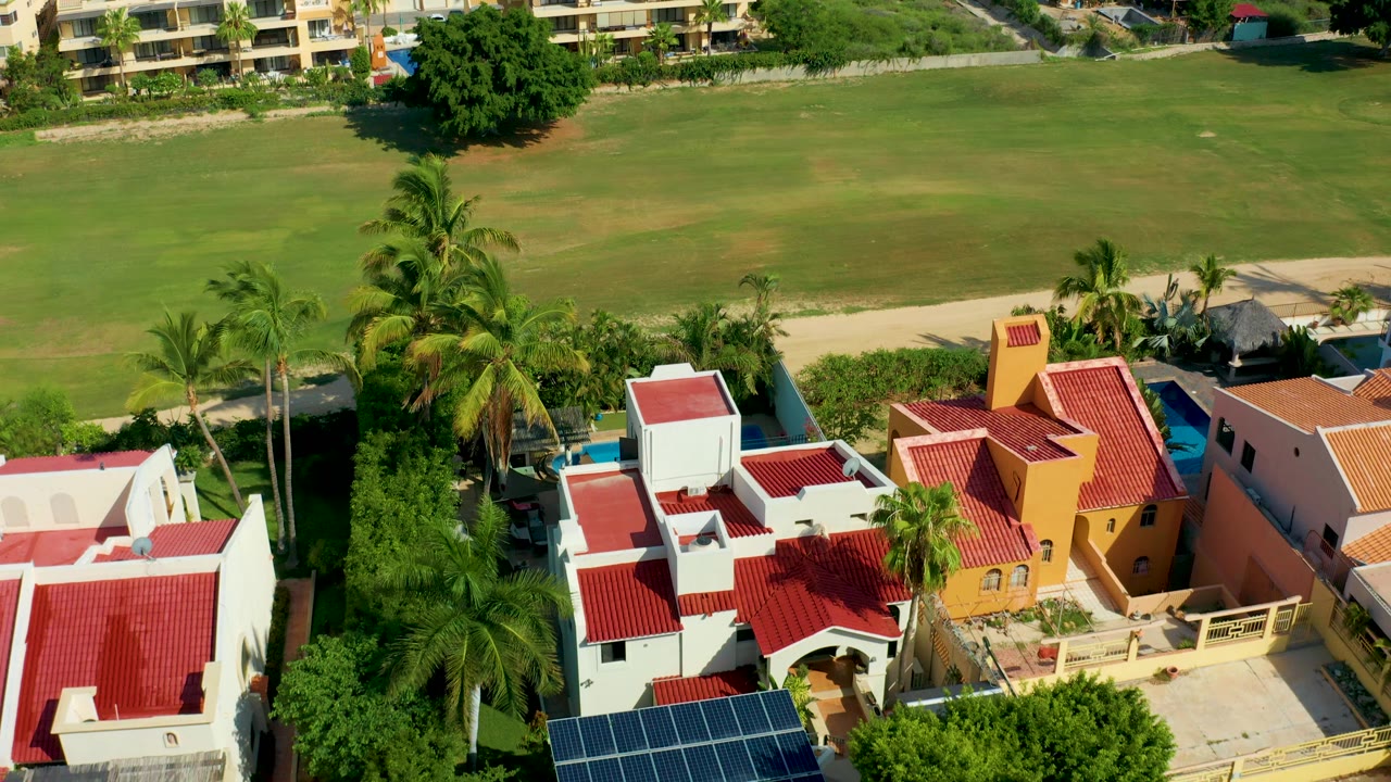 Aerial view in San Jose Del Cabo with red and orange rooflines, palm trees, a white villa, an ochre villa, driveways, and a grassy field behind, with no visible people.